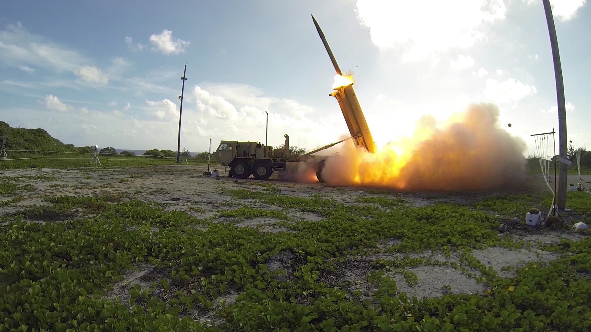 A THAAD interceptor launches into a blue daylight sky as bright exhaust and dust billow beside its launcher