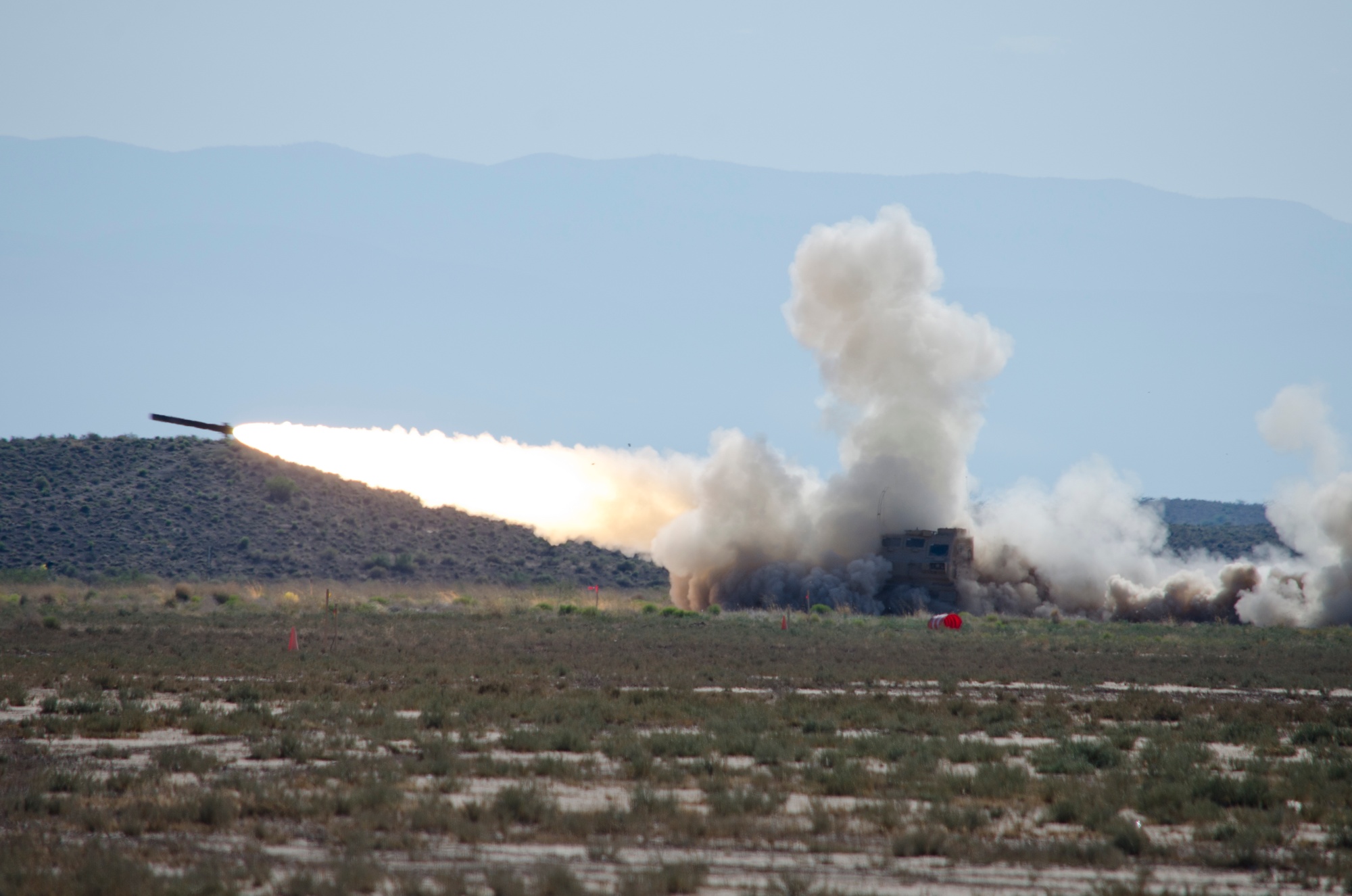 Rocket launching across a daylight test range, with a bright exhaust plume and gray dust against blue mountains